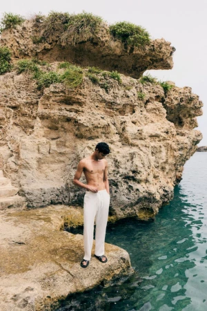 A dark-haired adult man stands on a rocky outcrop at the water's edge, wearing white trousers and sandals, with a rocky cliff behind.