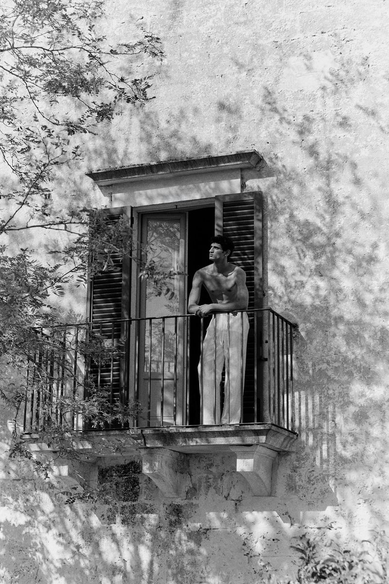 Black and white shot of a male model on a stone balcony with dark shutters, leaning and looking up with a serious expression.