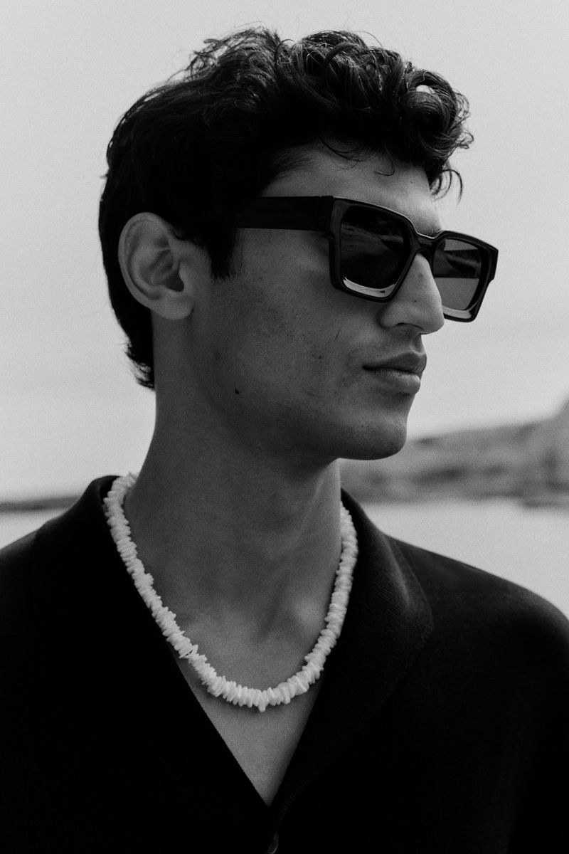 Portrait of a young man with curly hair and sunglasses, wearing a shell necklace against an outdoor background with water.