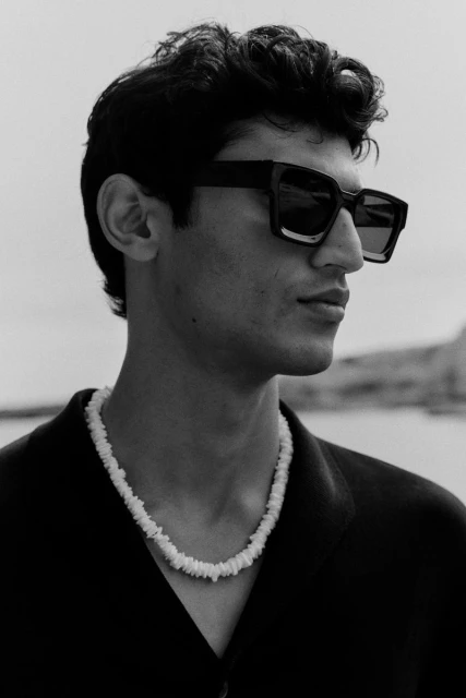 Portrait of a young man with curly hair and sunglasses, wearing a shell necklace against an outdoor background with water.