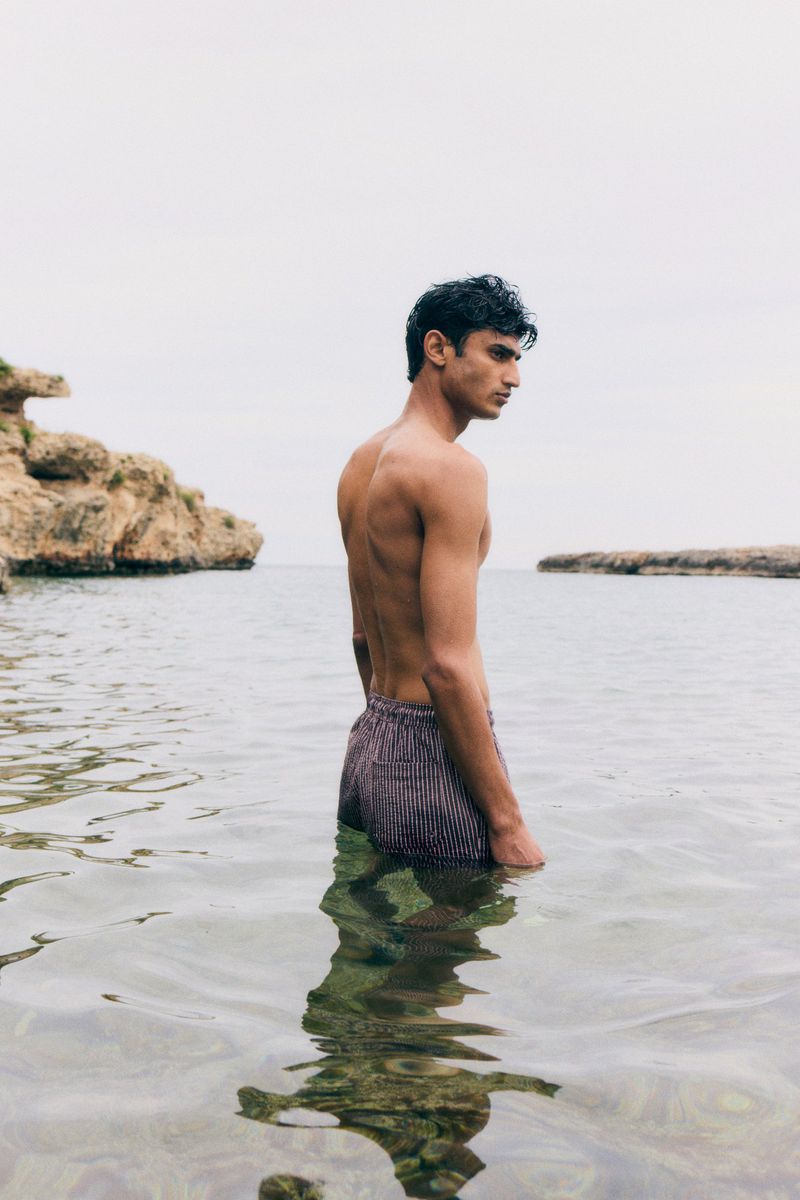 Young adult male stands in ocean water. The sky is overcast. Rocky landscape visible in the background. Portrait orientation.