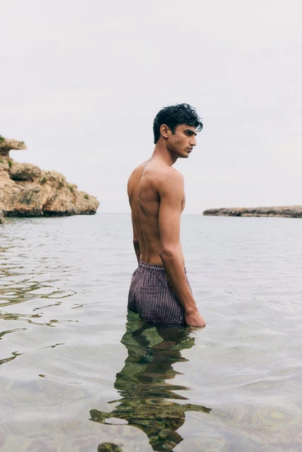 Young adult male stands in ocean water. The sky is overcast. Rocky landscape visible in the background. Portrait orientation.