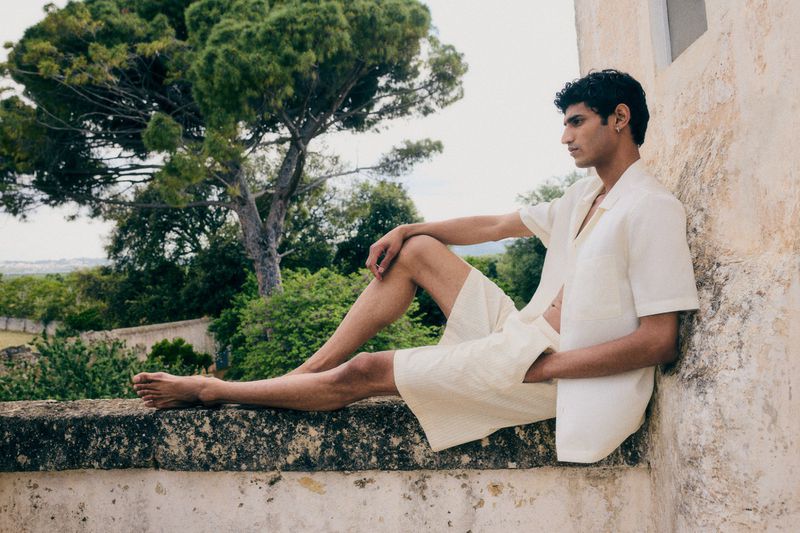 A young adult man in an off-white linen short set sits on a stone wall outside, surrounded by green foliage and trees in natural light.
