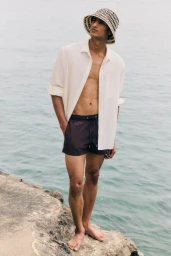 Model Neeraj Saini standing barefoot on a rocky shore wearing an unbuttoned white shirt, dark swim shorts, and a patterned bucket hat.