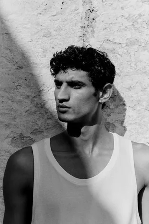 Model Neeraj Saini wearing a white tank top against a textured stone wall in high-contrast black and white.