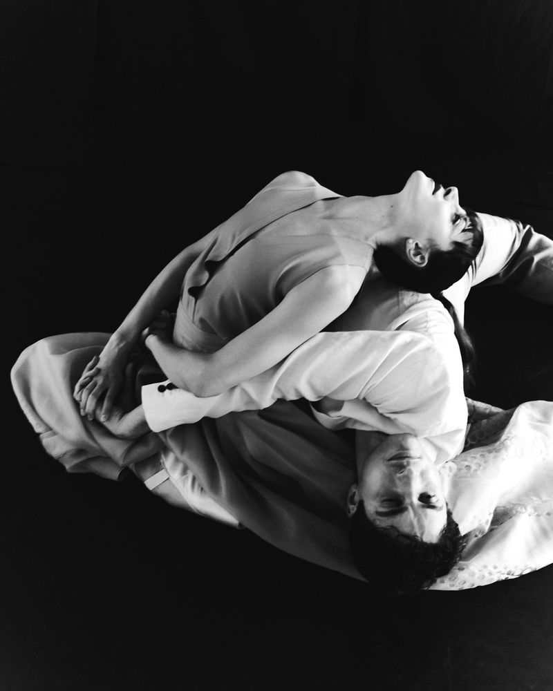 Monochrome artistic studio portrait of a man and woman, leaning towards each other with black background.