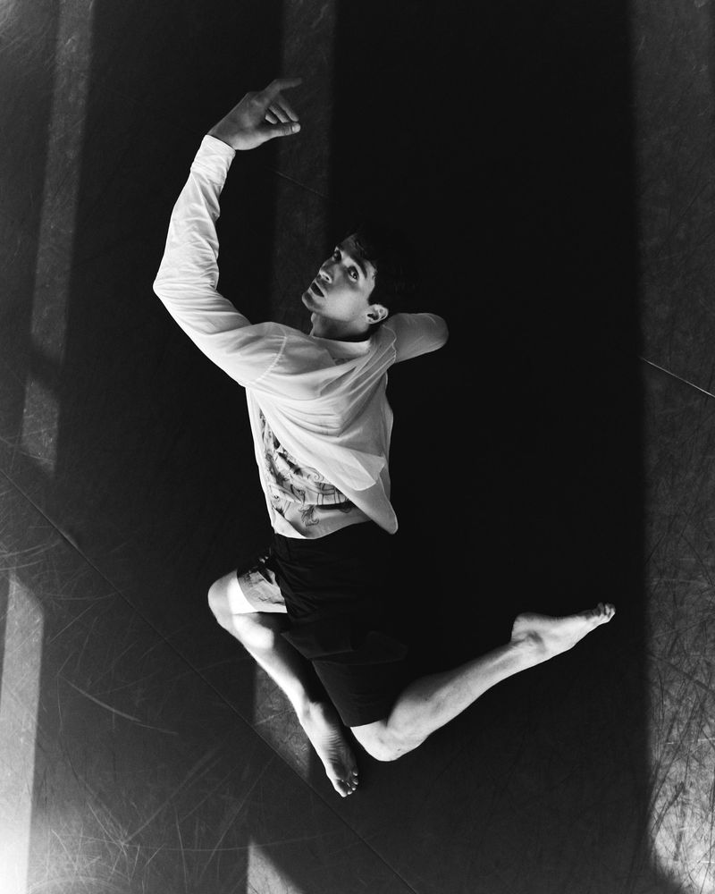 Monochrome shot of a young adult male leaping mid-air with arms raised, wearing a long-sleeve shirt and shorts, against a dark background.