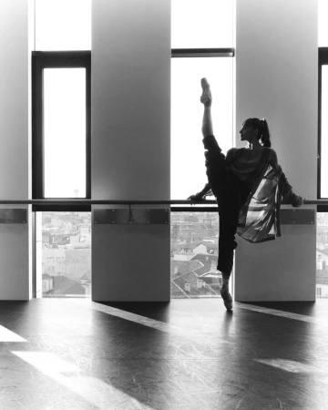 Black-and-white shot of a slender adult female dancer in elegant pose, wearing dance shoes, with a cityscape visible through windows.