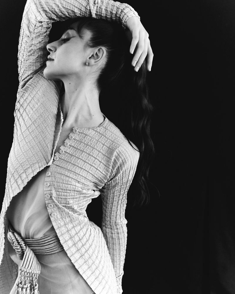 Monochrome portrait of a woman with hand behind her head, wearing a textured buttoned cardigan against a dark backdrop.