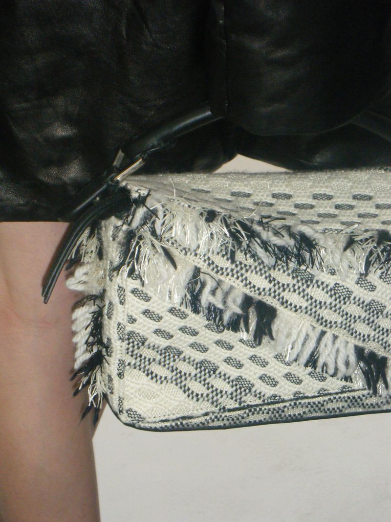 Close-up on a textured white clutch purse with black accents and fringe, held by a person in a black leather jacket.