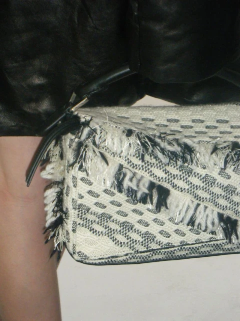 Close-up on a textured white clutch purse with black accents and fringe, held by a person in a black leather jacket.