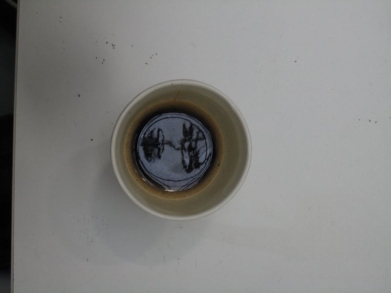 Top-down view of a tan ceramic mug filled with dark liquid, resting on a white surface with a few stray black particles nearby.