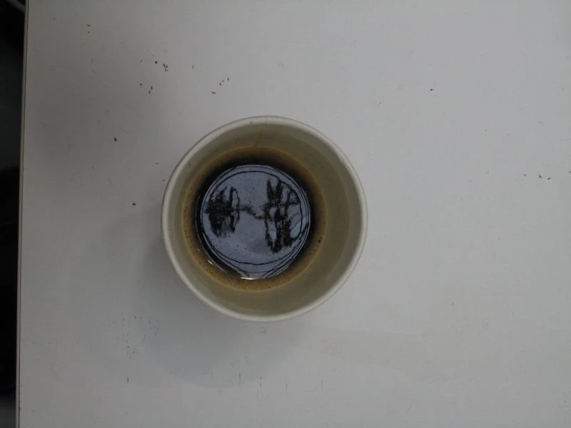 Top-down view of a tan ceramic mug filled with dark liquid, resting on a white surface with a few stray black particles nearby.