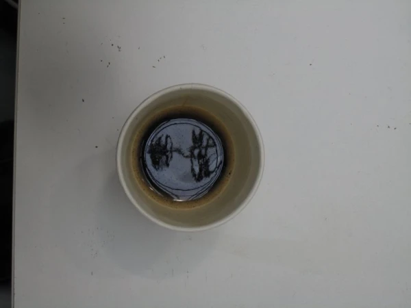 Top-down view of a tan ceramic mug filled with dark liquid, resting on a white surface with a few stray black particles nearby.