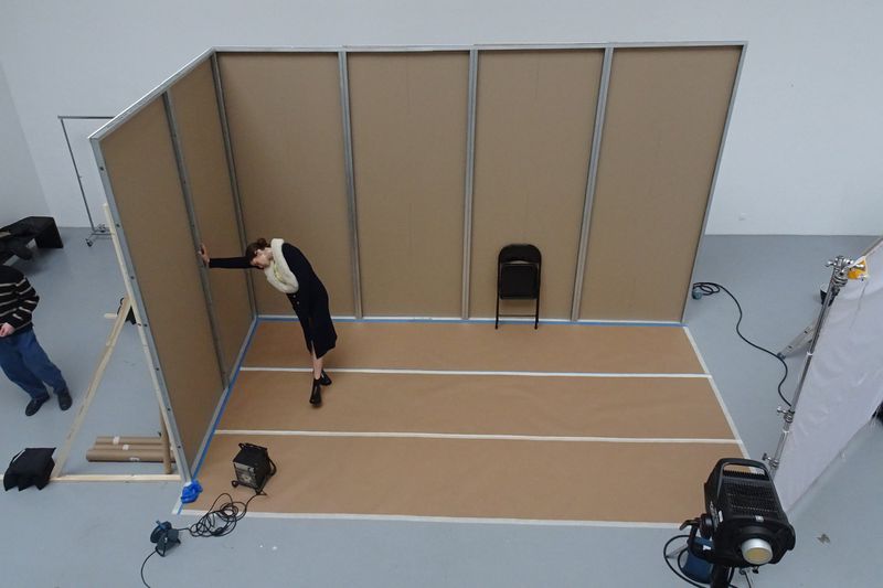 Woman in dark coat posing in a photography studio with grey walls and a brown floor with white lines, studio lighting, and equipment.