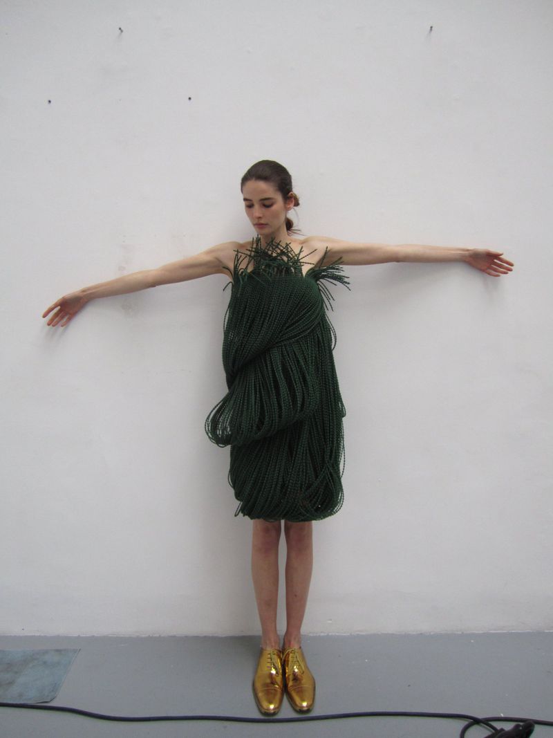 Full shot of a woman wearing a green rope dress and gold shoes, arms outstretched against a white wall, creating a unique fashion image.