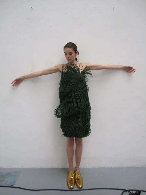 Full shot of a woman wearing a green rope dress and gold shoes, arms outstretched against a white wall, creating a unique fashion image.