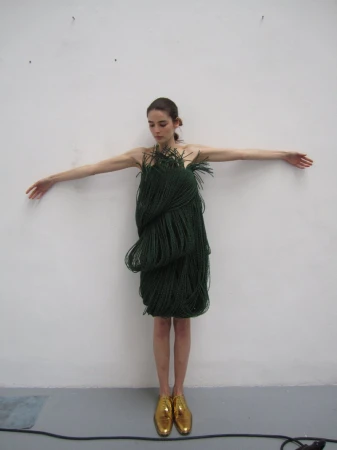 Full shot of a woman wearing a green rope dress and gold shoes, arms outstretched against a white wall, creating a unique fashion image.