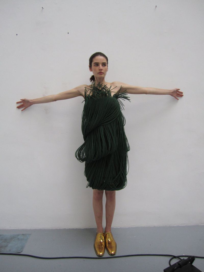 A woman in a dress made of dark green braided material stands with her arms outstretched against a white wall in golden shoes.