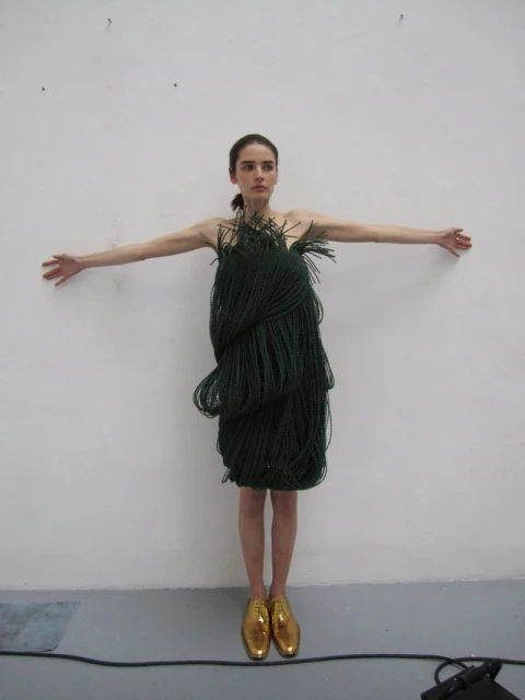 A woman in a dress made of dark green braided material stands with her arms outstretched against a white wall in golden shoes.