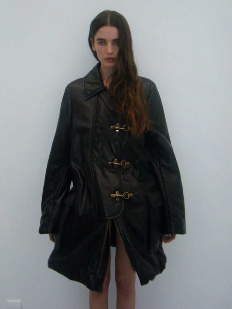 A woman poses in a black leather trench coat against a plain white background, highlighted by soft natural lighting.