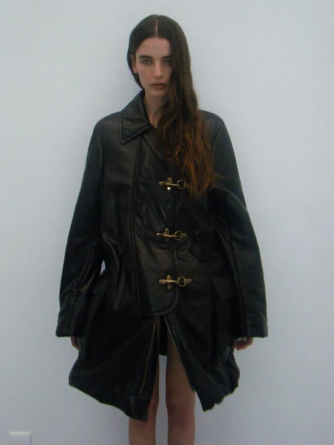 A woman poses in a black leather trench coat against a plain white background, highlighted by soft natural lighting.