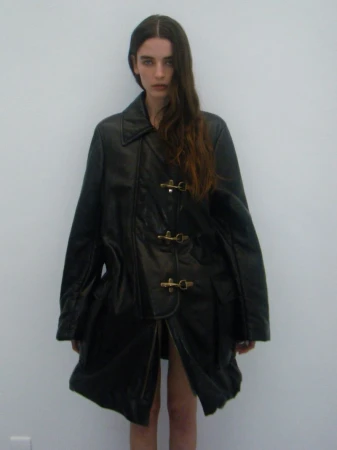 A woman poses in a black leather trench coat against a plain white background, highlighted by soft natural lighting.