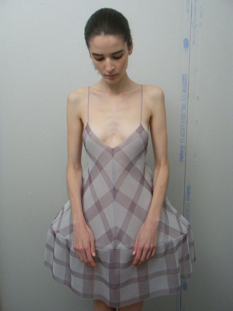 A woman in a short grey and burgundy plaid dress poses against a neutral background, looking downward with a contemplative expression.