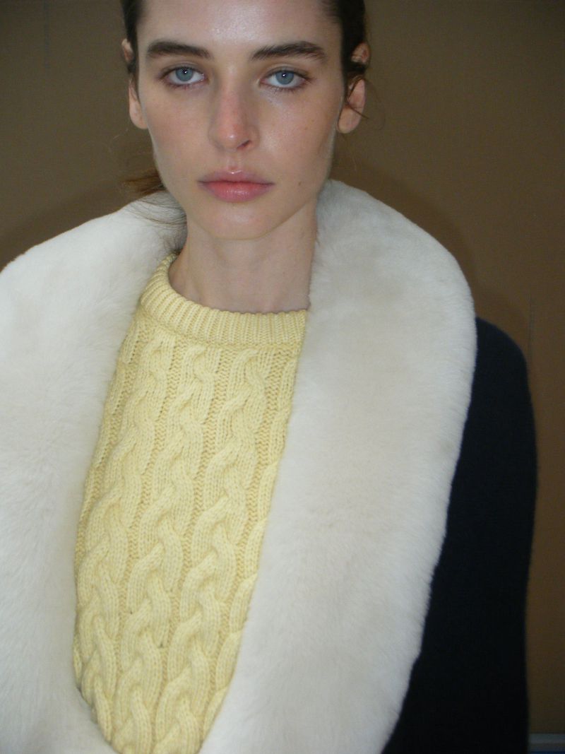 Close-up studio portrait of a fair-skinned woman with light blue eyes wearing a yellow cable-knit sweater and faux fur collar.