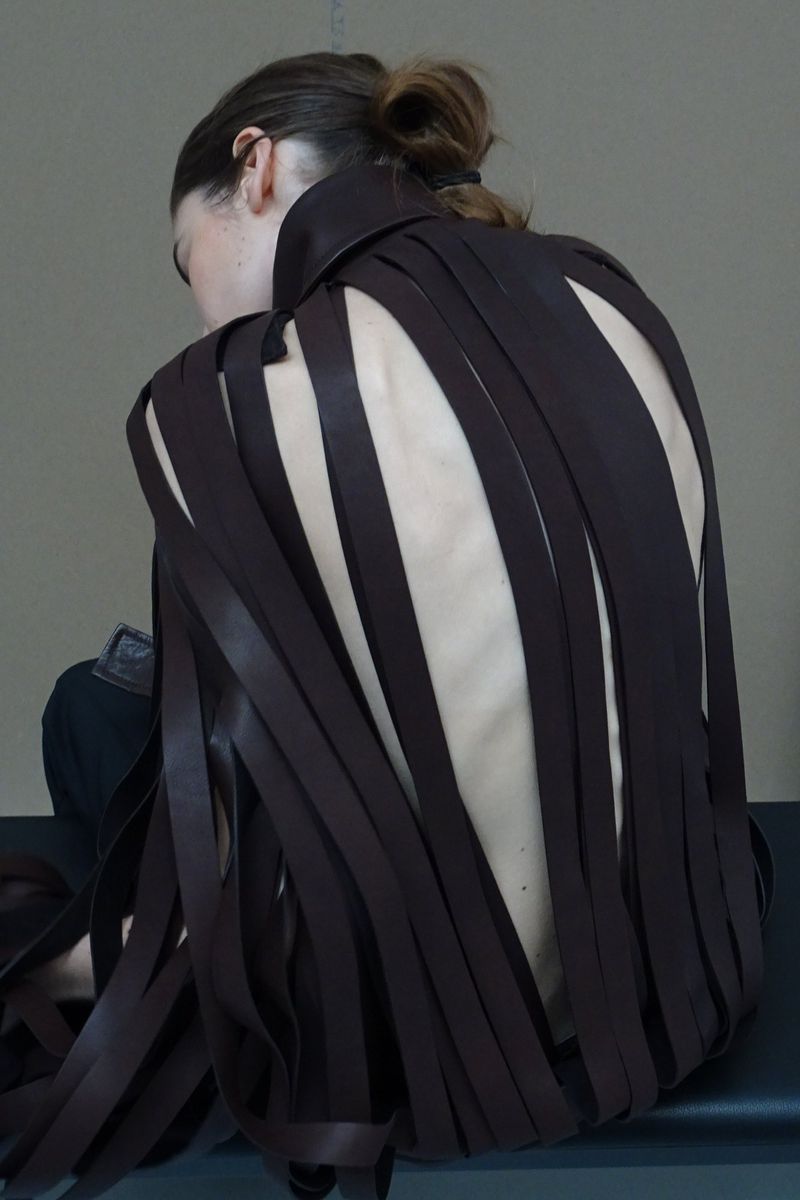 Back view of a woman with her hair in a bun, wearing a dark brown leather-strip top with open back, showcasing fashion and style.