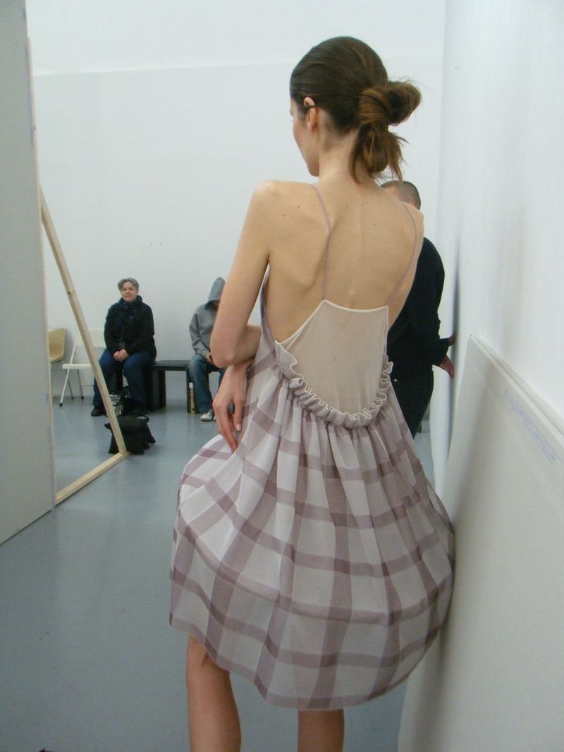 Back view of a fashion model in a plaid dress standing in a stark white studio with people sitting in the background.