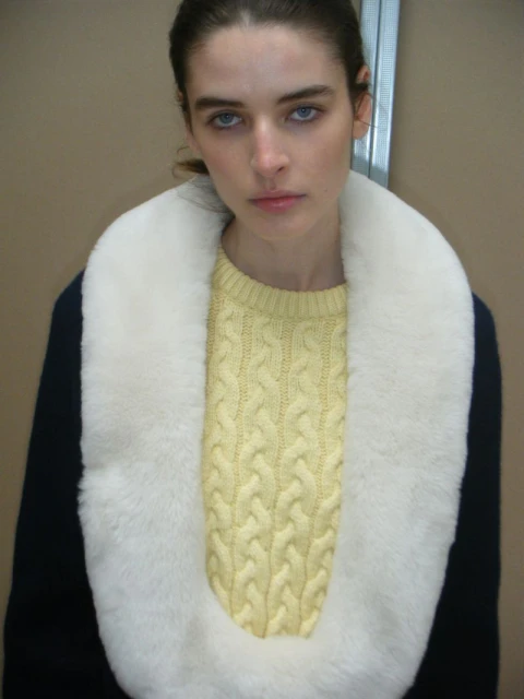 Close-up of a woman with fair skin, wearing a yellow sweater and white faux fur collar. Indoors, with neutral background and soft lighting.