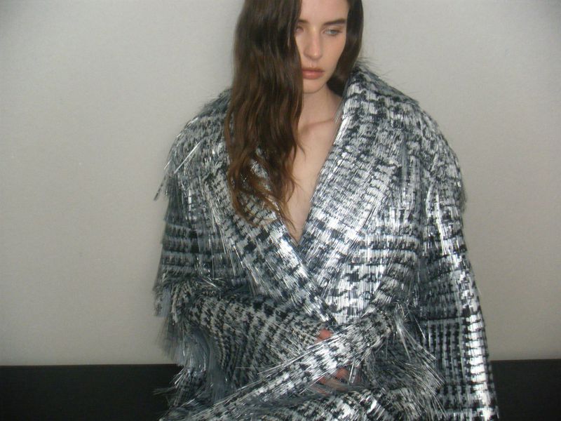 Woman in a shiny, checkered, fringe-trimmed coat, posing against a neutral wall; studio shot, fashion-forward aesthetic, and diffused lighting.