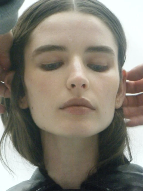 Close-up studio shot of an adult woman with brown hair being prepped by two hands, showcasing beauty or skincare in bright lighting.