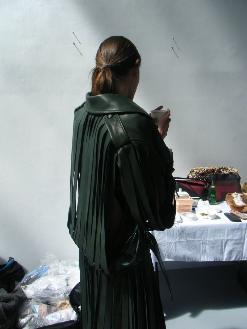 A woman stands indoors with her back to the camera, wearing a dark green fringe leather jacket and a pleated skirt with a ponytail.