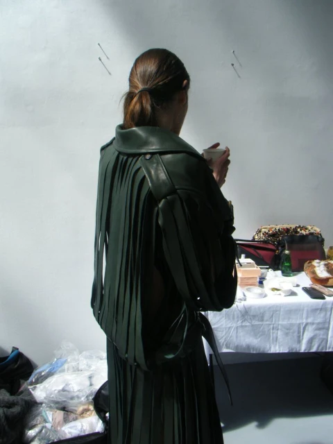 A woman stands indoors with her back to the camera, wearing a dark green fringe leather jacket and a pleated skirt with a ponytail.