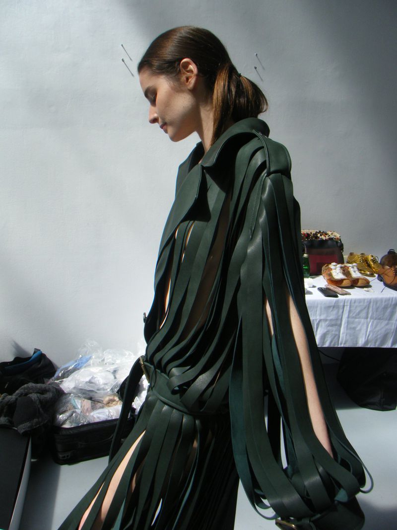 A woman poses in a green, fringe-sleeved fashion coat in a photo studio with a white wall backdrop and miscellaneous objects on a table.