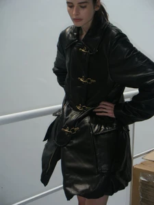 Model Libby Taverner wearing an oversized black leather coat with gold toggle closures beside a white railing.