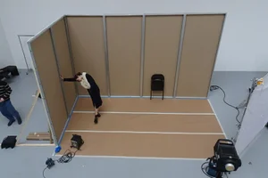 High-angle studio view of a model in a dark dress posing against brown cardboard set walls during a fashion photoshoot.