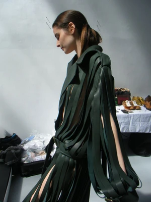 Model Libby Taverner in a dark green slotted fringe coat looking downward backstage during a fashion shoot.