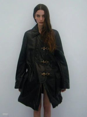 Model Libby Taverner wearing an oversized black leather coat with gold toggle clasps against a plain white studio background.