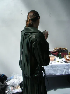 Model viewed from behind wearing a dark green fringed leather jacket and holding a small cup against a white wall.
