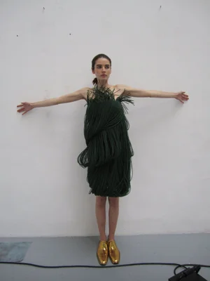Model in a dark green fringed dress with arms outstretched, wearing gold shoes against a neutral backdrop