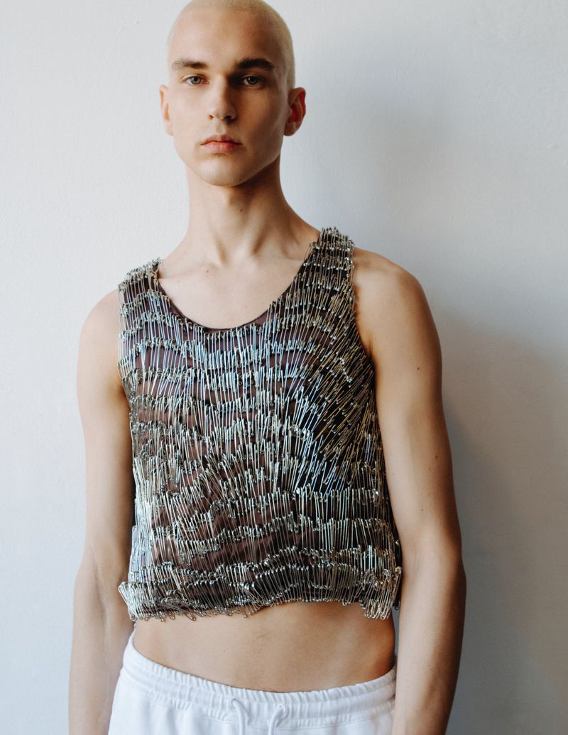 Person wearing a tank top made of safety pins posing against a white background, cropped torso shot.