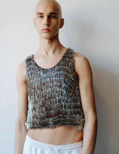 Person wearing a tank top made of safety pins posing against a white background, cropped torso shot.