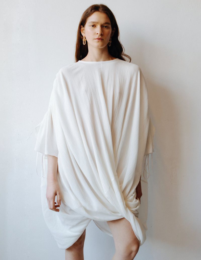 Model Mica Tosi wearing an oversized draped off-white dress and gold drop earrings against a neutral studio backdrop.