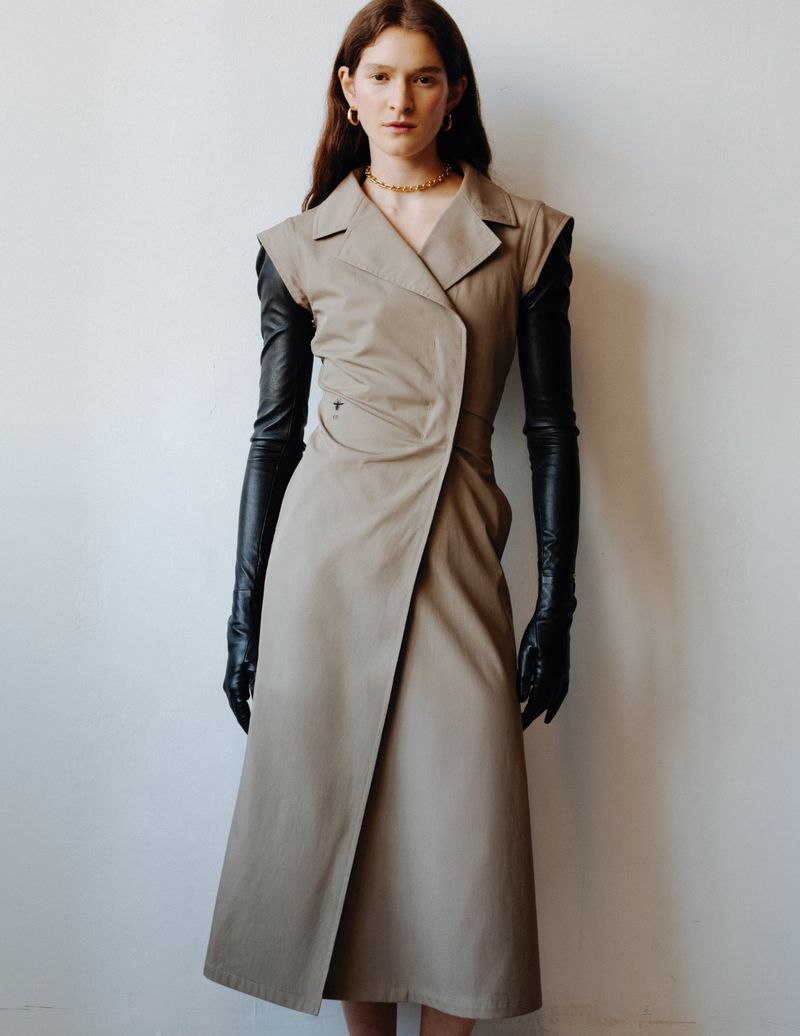 A woman stands indoors wearing a sleeveless trench dress and long black gloves, looking forward at the camera with neutral expression.