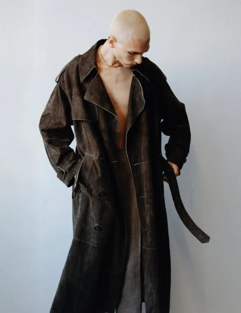 A bald man with fair skin is wearing a brown trench coat and a gold chain against a neutral wall, looking down with a subdued expression.