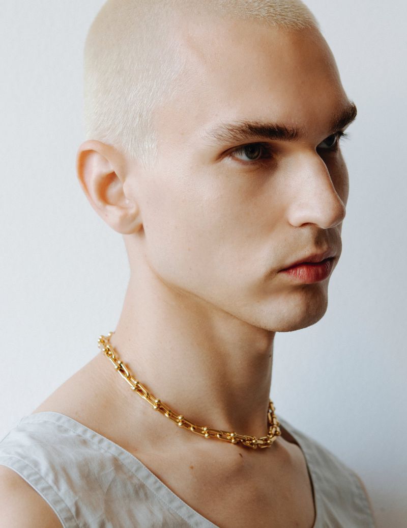 Close-up portrait of a young adult male with platinum blonde buzzcut wearing a gold necklace against a white background.