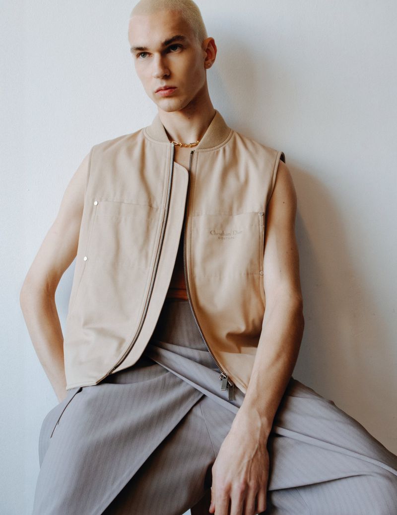 Portrait of an adult male model wearing a tan vest and grey trousers, sitting indoors against a plain white wall in soft light.